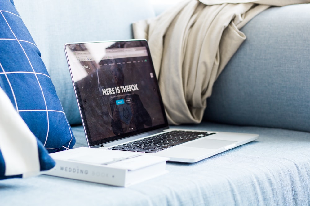 A laptop sitting next to a book on a couch.