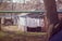Clothes are drying on a line outdoors in a rustic backyard setting. The area is surrounded by makeshift structures and leafless trees, indicating a rural or underdeveloped location. The foreground features grass and bare patches of ground, while the background includes a wooden shed and older buildings.