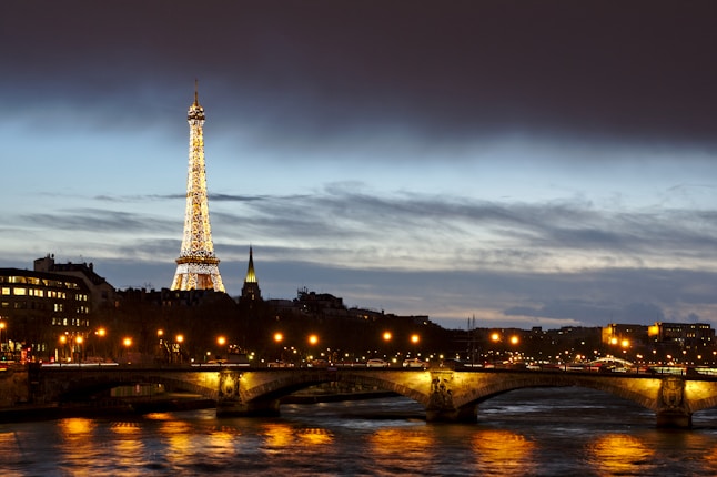 A beautifully illuminated Eiffel Tower stands prominently against the evening sky, surrounded by city lights reflecting off the Seine River. The clouds add depth to the twilight atmosphere, while a bridge with glowing street lamps spans the river.