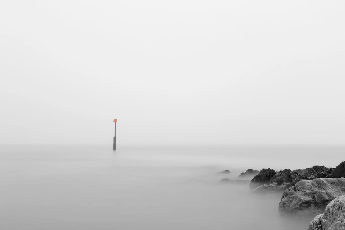 A thin channel marker standing in flat, misty lake water, with dark rocks at the lower right