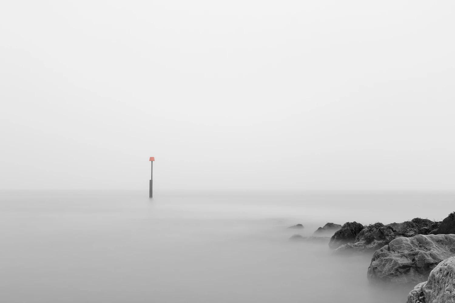 A thin channel marker standing in flat, misty lake water, with dark rocks at the lower right