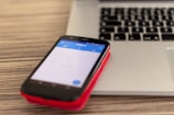 A smartphone with a visible Skype app screen is placed on a red case, resting on top of a closed laptop on a wooden surface. The laptop has a black keyboard and a silver body.