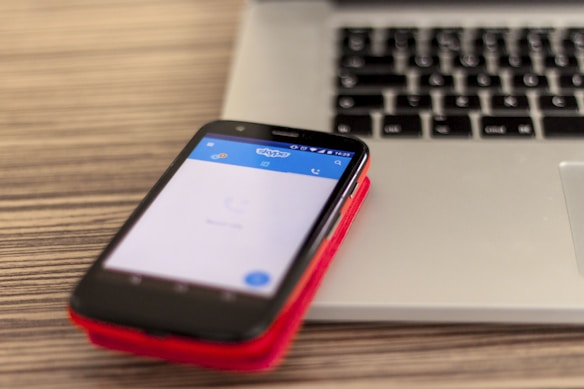 A smartphone with a visible Skype app screen is placed on a red case, resting on top of a closed laptop on a wooden surface. The laptop has a black keyboard and a silver body.