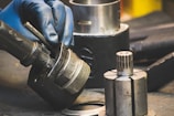 A gloved hand holding and assembling or disassembling a metallic component, possibly part of machinery or an engine. Several other cylindrical metallic parts and tools are situated around a workstation.