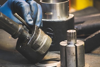 A gloved hand holding and assembling or disassembling a metallic component, possibly part of machinery or an engine. Several other cylindrical metallic parts and tools are situated around a workstation.