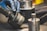 A gloved hand holding and assembling or disassembling a metallic component, possibly part of machinery or an engine. Several other cylindrical metallic parts and tools are situated around a workstation.