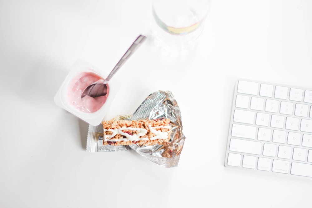 A spoon inside a small yogurt cup next to a granola bar and a Mac keyboard.