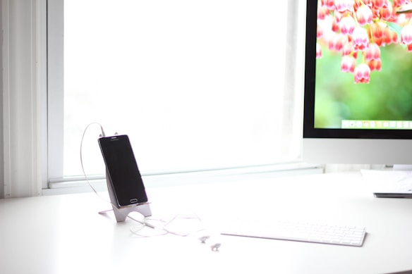 A sleek and modern workspace featuring a smartphone on a stand with earphones attached, placed on a white desk. A desktop computer with a vibrant floral wallpaper is positioned nearby. The room is well-lit, with large windows allowing natural light to flood the space.