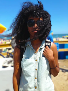 A person stands on a sunny beach, wearing sunglasses and a light denim button-up shirt. Their hair is windblown, and they hold onto a backpack strap. The background is blurred but shows a bright sky and some indistinct beach elements.