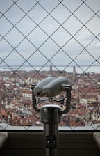 A scenic view of a historic city with red-tiled roofs and distant towers, observed through a wire-mesh fence. In the foreground, a metallic binocular viewer is prominently positioned, inviting viewers to explore the cityscape.