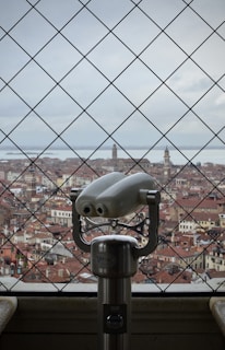 A scenic view of a historic city with red-tiled roofs and distant towers, observed through a wire-mesh fence. In the foreground, a metallic binocular viewer is prominently positioned, inviting viewers to explore the cityscape.