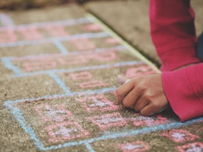 A child wearing a red long-sleeve shirt draws with colored chalk on a sidewalk, creating a hopscotch grid with numbers using pink and blue colors.
