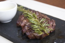 A succulent piece of grilled steak garnished with a sprig of fresh rosemary is placed on a dark slate platter. Beside the steak, there is a small white bowl filled with a creamy sauce.