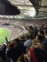A crowded stadium filled with spectators during a sports event. The audience is animated, with some individuals holding signs and others clapping. The stadium is well-lit with a clear view of the field on the left side. The architecture of the stadium has a modern, open-roof design.