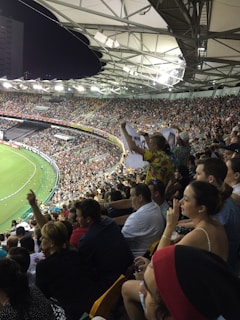 A crowded stadium filled with spectators during a sports event. The audience is animated, with some individuals holding signs and others clapping. The stadium is well-lit with a clear view of the field on the left side. The architecture of the stadium has a modern, open-roof design.