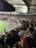 A crowded stadium filled with spectators during a sports event. The audience is animated, with some individuals holding signs and others clapping. The stadium is well-lit with a clear view of the field on the left side. The architecture of the stadium has a modern, open-roof design.