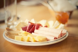 A plate with an assortment of deli meats, cheese slices, and crackers. Some of the meats are rolled, and there is a blurry background featuring a glass and a jar of preserves.