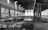An abandoned industrial building with a large open space featuring metal beams and a broken floor. The walls and surfaces are covered with various graffiti and tags. Large windows allow light to filter in from the right side, casting shadows and creating a contrast with the dark interior.