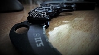 A close-up view of a curved, textured knife with a black handle sitting on a wooden surface. The blade is partially visible, showcasing the logo and sharp edges under dim lighting.