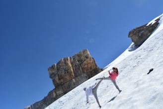 Two people on a snowy slope practicing martial arts or performing a high kick. The scene includes a large rock formation in the background, and the sky is clear and bright blue.