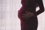A pregnant person is gently cradling their stomach with both hands, wearing a dark maroon dress. The background is softly lit with a sheer curtain, creating a serene and intimate atmosphere.