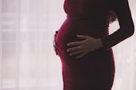 A pregnant person is gently cradling their stomach with both hands, wearing a dark maroon dress. The background is softly lit with a sheer curtain, creating a serene and intimate atmosphere.