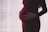 A pregnant person is gently cradling their stomach with both hands, wearing a dark maroon dress. The background is softly lit with a sheer curtain, creating a serene and intimate atmosphere.