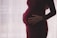 A pregnant person is gently cradling their stomach with both hands, wearing a dark maroon dress. The background is softly lit with a sheer curtain, creating a serene and intimate atmosphere.