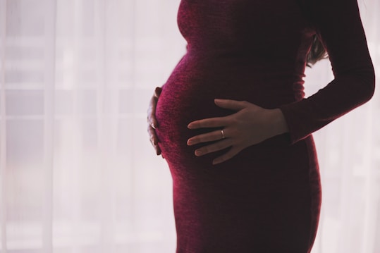A pregnant person is gently cradling their stomach with both hands, wearing a dark maroon dress. The background is softly lit with a sheer curtain, creating a serene and intimate atmosphere.