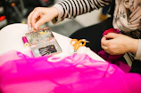 A person is engaged in sewing or tailoring, with their hands working with a piece of pink fabric. Various sewing tools are on the table, including a box filled with colorful pins, a spool of pink thread, and a pair of small, orange-handled scissors. The person is using a thimble on their finger.