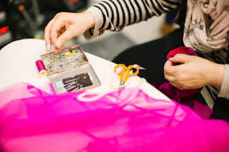 A person is engaged in sewing or tailoring, with their hands working with a piece of pink fabric. Various sewing tools are on the table, including a box filled with colorful pins, a spool of pink thread, and a pair of small, orange-handled scissors. The person is using a thimble on their finger.