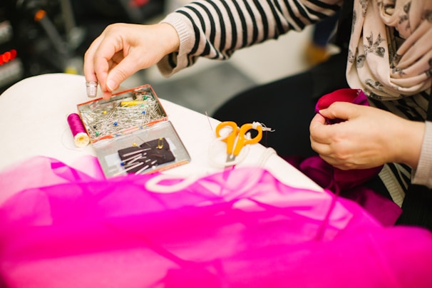 A person is engaged in sewing or tailoring, with their hands working with a piece of pink fabric. Various sewing tools are on the table, including a box filled with colorful pins, a spool of pink thread, and a pair of small, orange-handled scissors. The person is using a thimble on their finger.