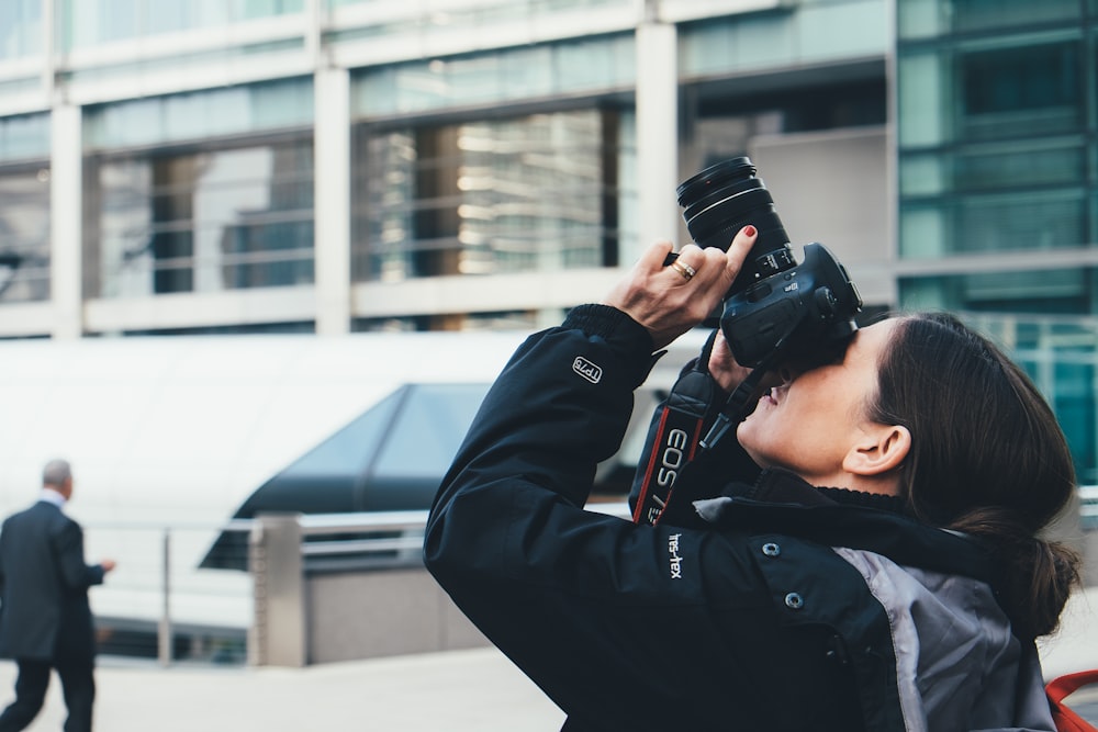 femme portant une veste grise et noire à l’aide d’un appareil photo reflex numérique