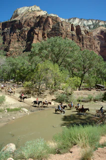 Horseback riders crossing a wild river with mountains looming in the background.