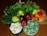 A variety of fresh fruits and vegetables are arranged on a wooden surface. There are red apples, green pears, vibrant green leafy vegetables, orange and red bell peppers, and bowls containing broccoli, cauliflower, and green beans.