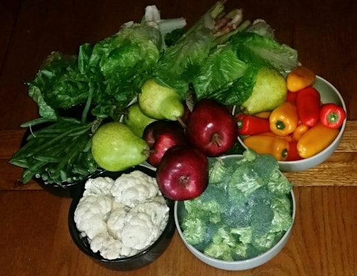 A variety of fresh fruits and vegetables are arranged on a wooden surface. There are red apples, green pears, vibrant green leafy vegetables, orange and red bell peppers, and bowls containing broccoli, cauliflower, and green beans.