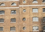 A brick building facade featuring multiple windows with a variety of shapes including arched and circular designs. The building has a traditional architectural style with barred windows and a uniform brick pattern.