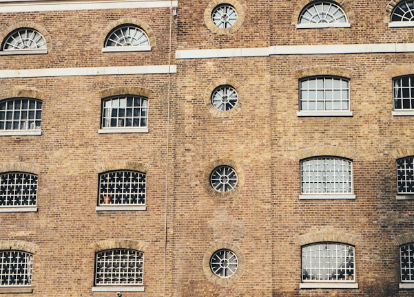 A brick building facade featuring multiple windows with a variety of shapes including arched and circular designs. The building has a traditional architectural style with barred windows and a uniform brick pattern.