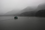 A serene landscape features a dark, misty lake surrounded by mountains shrouded in fog. A small green house floats on the water, connected to a narrow wooden pathway. The surrounding forest blends into the mist, creating a tranquil and mysterious atmosphere.