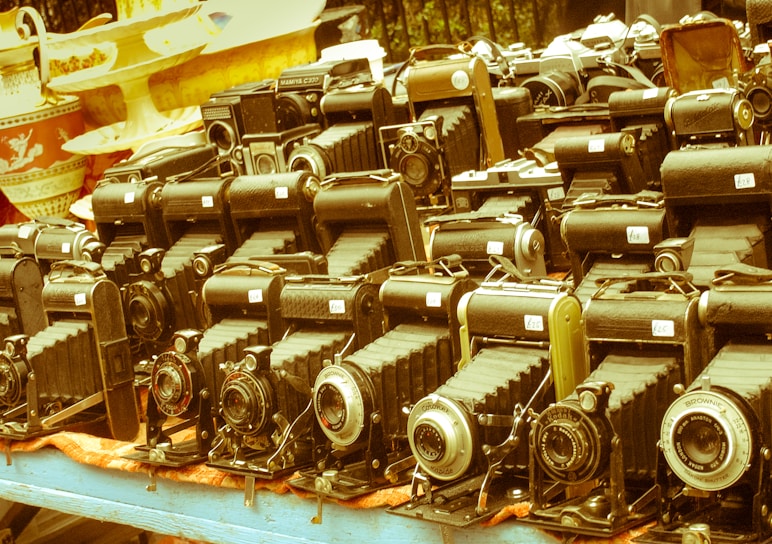 A collection of vintage cameras displayed on a table suggests an antique or flea market setting. The cameras have a classic bellows design and are arranged in several neat rows. Numerous price tags are visible on each camera, indicating they are for sale. The background shows decorative ceramic dishes, adding to the nostalgic atmosphere.