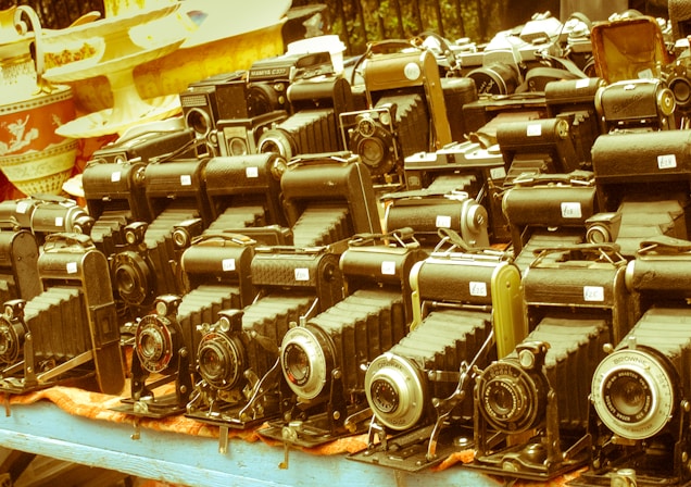 A collection of vintage cameras displayed on a table suggests an antique or flea market setting. The cameras have a classic bellows design and are arranged in several neat rows. Numerous price tags are visible on each camera, indicating they are for sale. The background shows decorative ceramic dishes, adding to the nostalgic atmosphere.