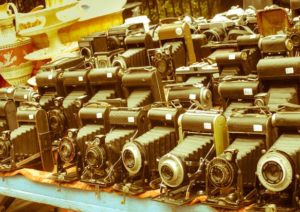 A collection of vintage cameras displayed on a table suggests an antique or flea market setting. The cameras have a classic bellows design and are arranged in several neat rows. Numerous price tags are visible on each camera, indicating they are for sale. The background shows decorative ceramic dishes, adding to the nostalgic atmosphere.