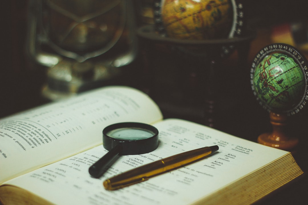 A book with a magnifying glass on top of it, next to a pen, and globes on a desk in Cianorte