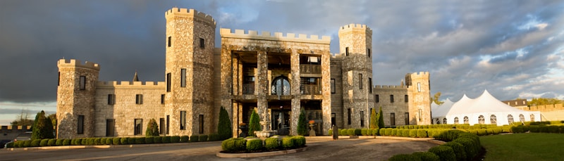 A large stone castle-like building with square towers and crenellated walls is set under a partly cloudy sky. The foreground features a circular driveway with a fountain surrounded by neatly trimmed hedges and shrubs. To the side, a large white tent with arch-top windows stands on the grass, indicating a possible event setting.