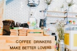 An arrangement of coffee-related items is displayed, including a variety of cups, cutlery, and a coffee grinder in a bright and inviting café setting. A humorous sign reads 'COFFEE DRINKERS MAKE BETTER LOVERS'. The setting features white brick walls, shelves lined with colorful mugs and jars, and decorative plants, creating a cozy and lively atmosphere.