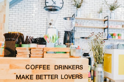 An arrangement of coffee-related items is displayed, including a variety of cups, cutlery, and a coffee grinder in a bright and inviting café setting. A humorous sign reads 'COFFEE DRINKERS MAKE BETTER LOVERS'. The setting features white brick walls, shelves lined with colorful mugs and jars, and decorative plants, creating a cozy and lively atmosphere.