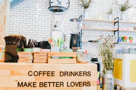 An arrangement of coffee-related items is displayed, including a variety of cups, cutlery, and a coffee grinder in a bright and inviting caf&eacute; setting. A humorous sign reads 'COFFEE DRINKERS MAKE BETTER LOVERS'. The setting features white brick walls, shelves lined with colorful mugs and jars, and decorative plants, creating a cozy and lively atmosphere.