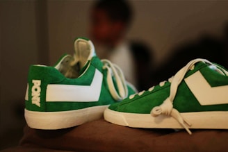 A pair of green and white sneakers positioned on a surface. The sneakers have a sporty design with the brand name 'Pony' visible on the heel of one shoe. The background is blurred, and there is a person in the background, out of focus.