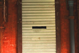 A closed metal roller shutter with horizontal ridges set within a reddish brick wall. A small rectangular cut-out is visible in the center of the door. The wall has a pipe running vertically on the right side.