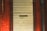 A closed metal roller shutter with horizontal ridges set within a reddish brick wall. A small rectangular cut-out is visible in the center of the door. The wall has a pipe running vertically on the right side.